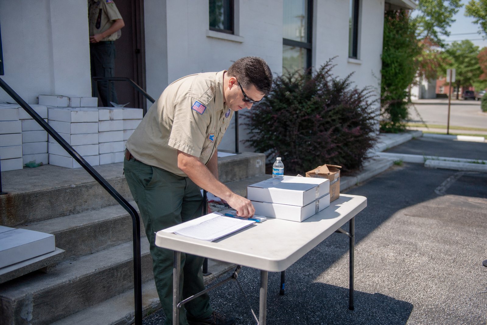 A troop leader organizing stacked cookie cases and a paper signup sheet on a folding table outside a community building